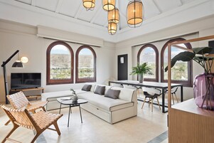 A modern living room with a sofa, chairs, a dining table, and large arched windows offering a view of the landscape.