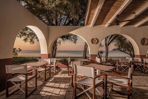 Un área de comedor al aire libre con mesas y sillas, amplias ventanas en arco y vista al mar.