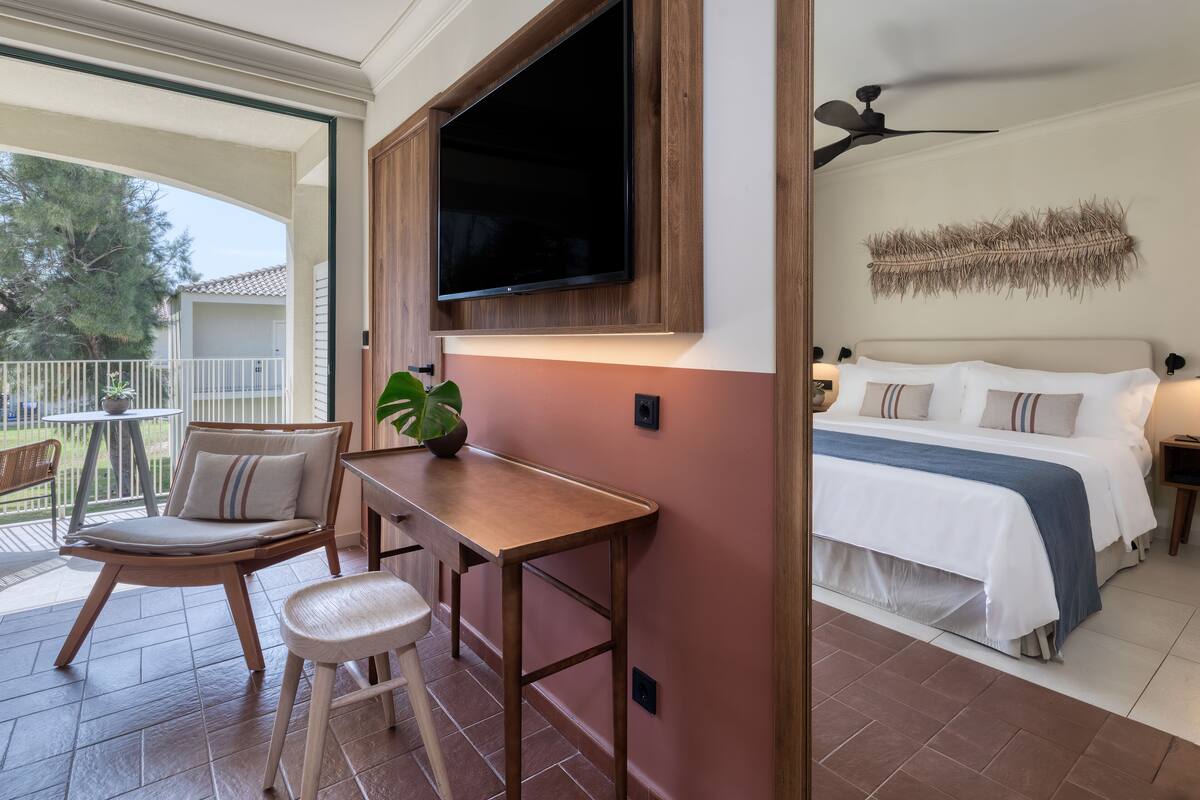A hotel room with a bed, a desk with a chair, a TV mounted on the wall, and a balcony with a chair and a plant.
