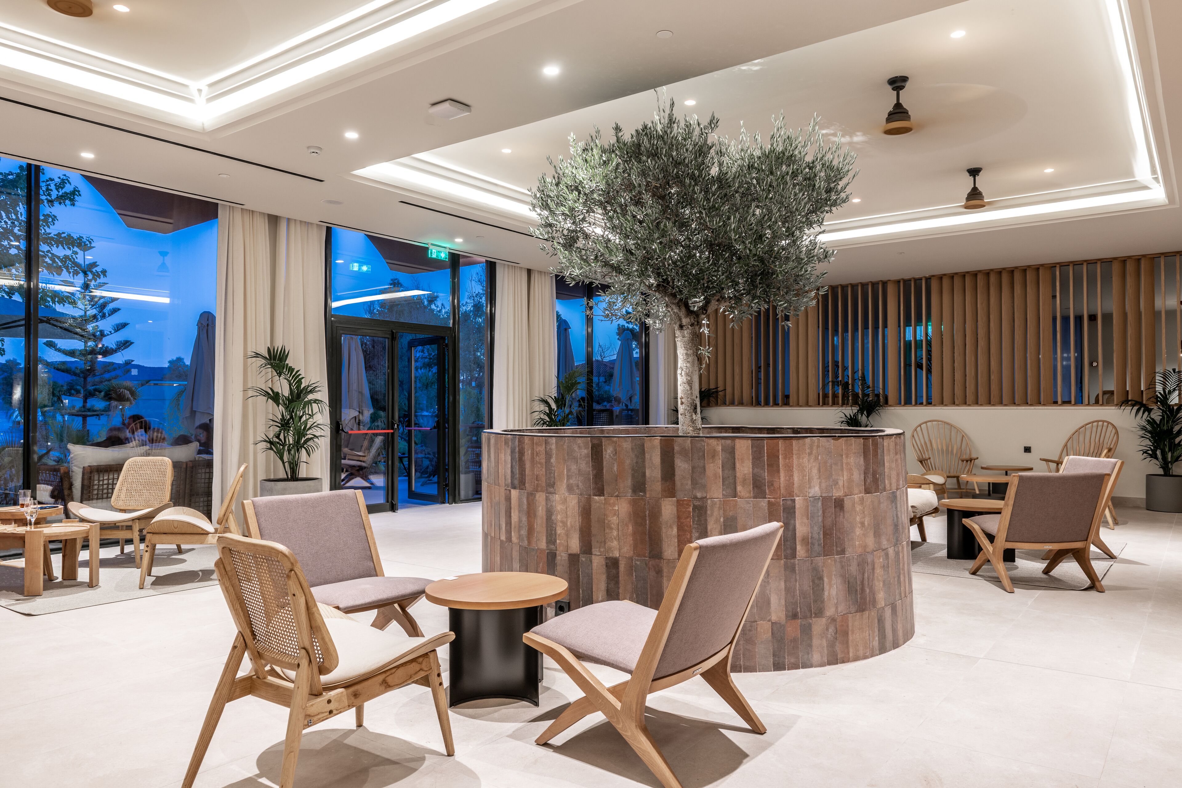 A modern lobby with wooden furniture, a central plant, and large windows offering a view of the outdoors.