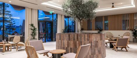 A modern lobby with wooden furniture, a central plant, and large windows offering a view of the outdoors.