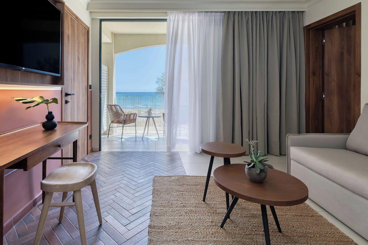 A modern hotel room with a wooden desk, two stools, a sofa, and a TV. There is a balcony with a view of the sea.