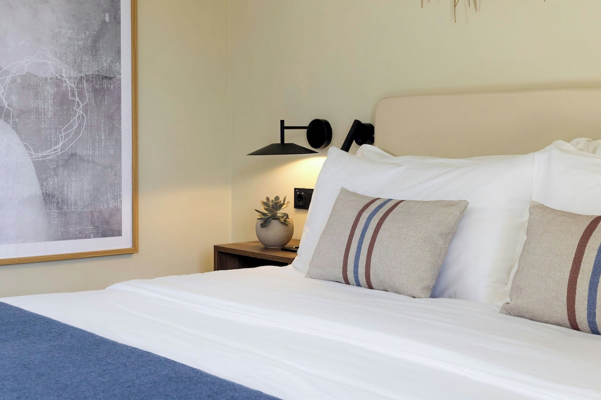 A neatly made bed with white linens and striped pillows, a bedside lamp, and a framed abstract painting on the wall.