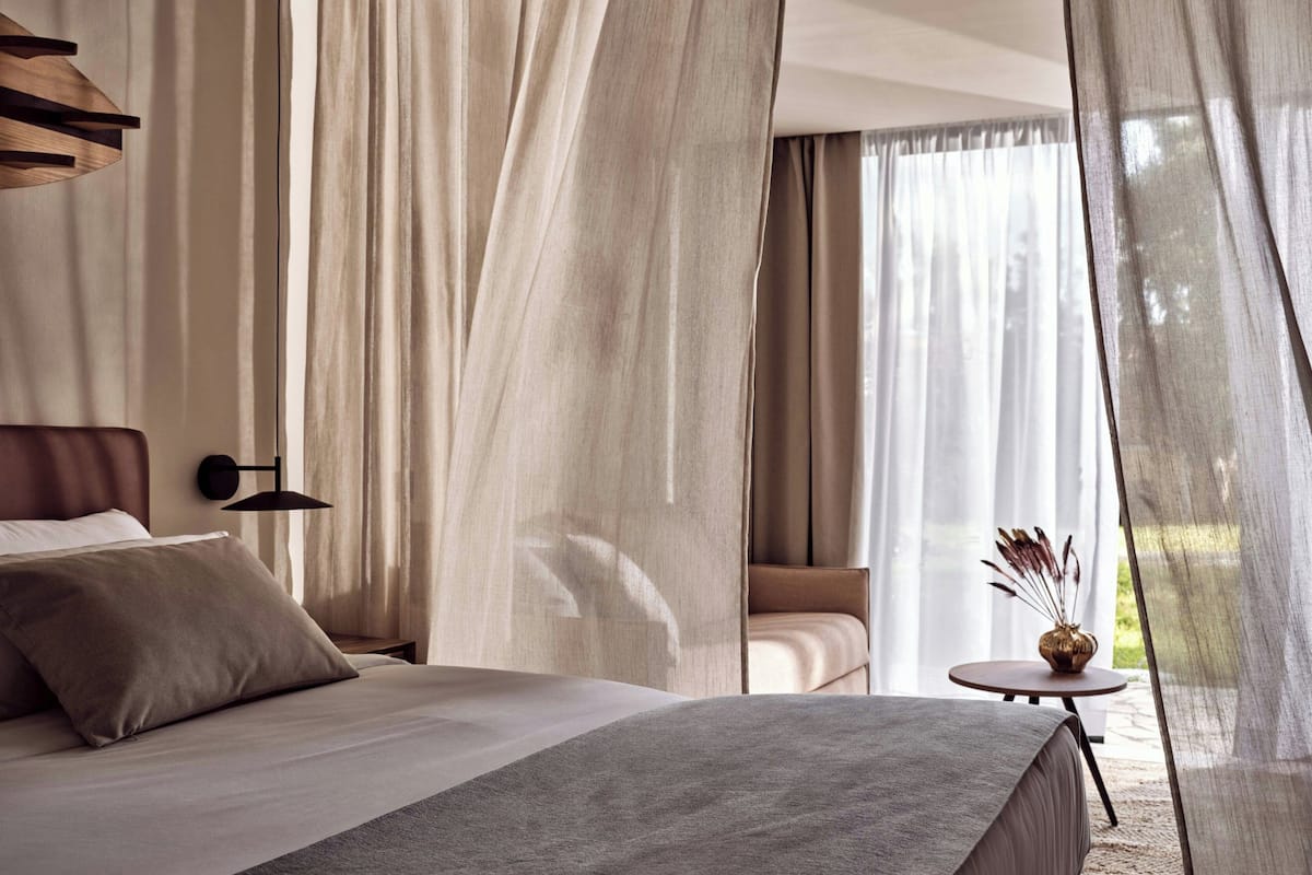 A modern bedroom with a large bed, a bedside table, a small round table with a vase, and sheer curtains.