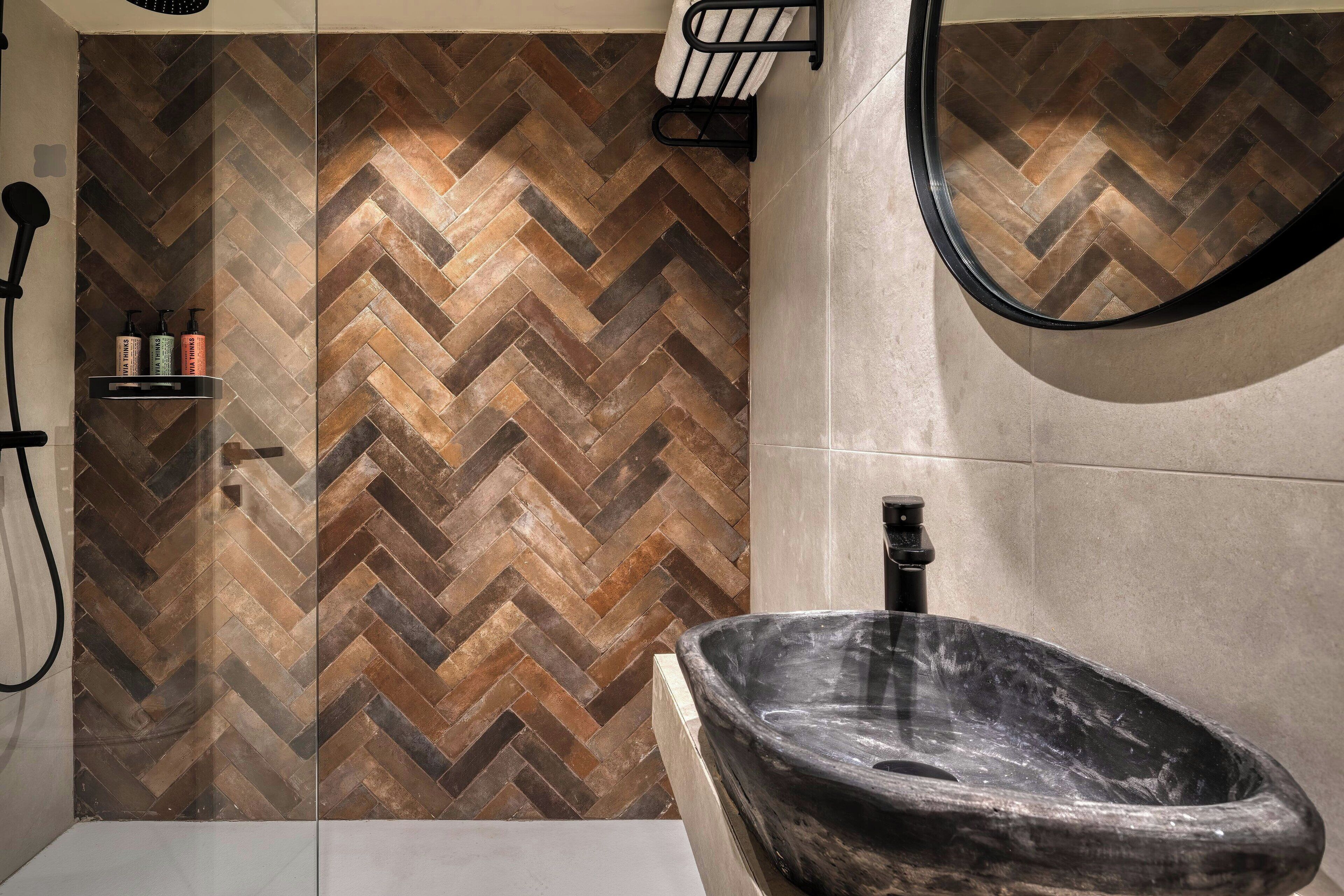 A bathroom with a herringbone-patterned tile wall, a freestanding stone sink, and a shower area with a glass door.