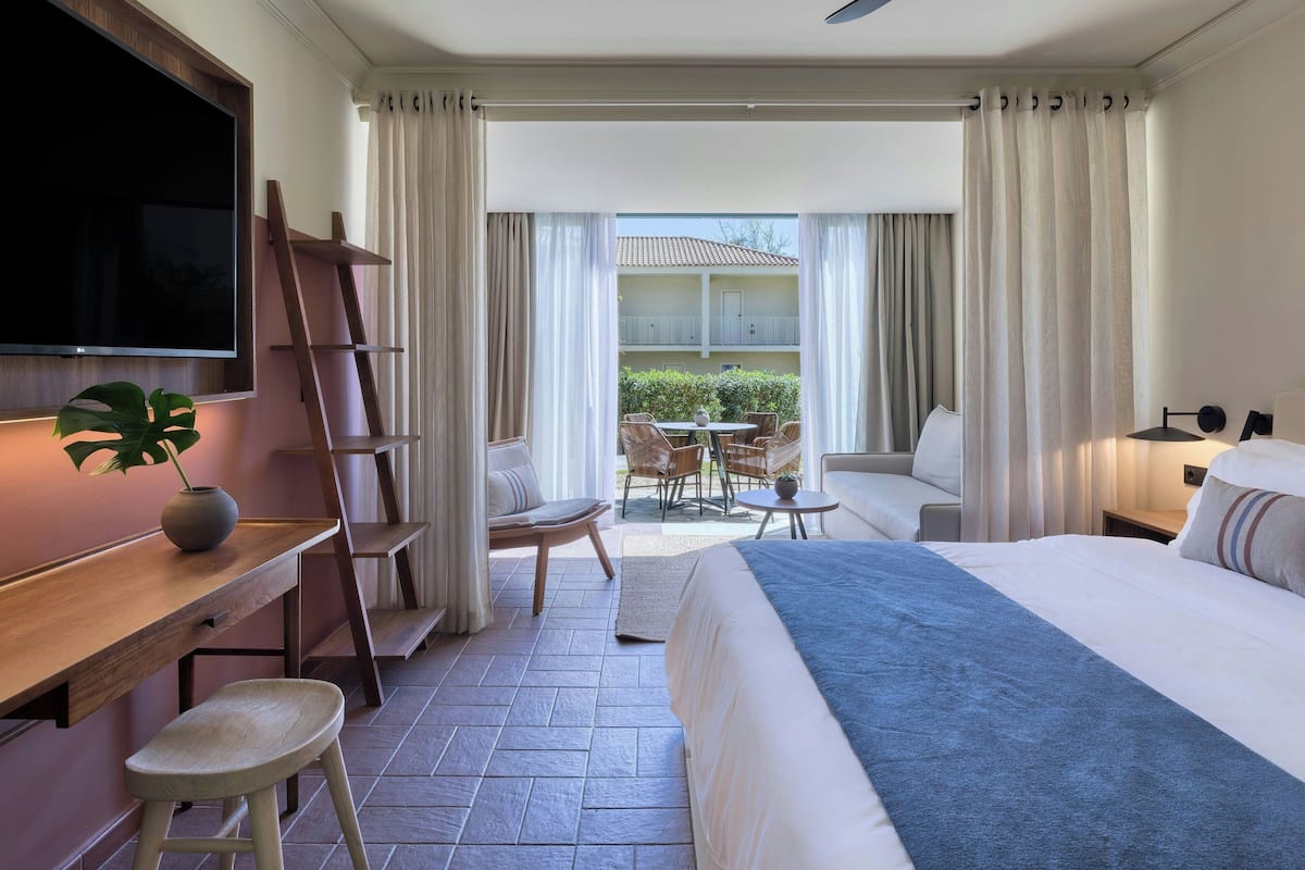 A hotel room with a bed, a desk with a chair, a TV mounted on the wall, and a balcony with outdoor seating.
