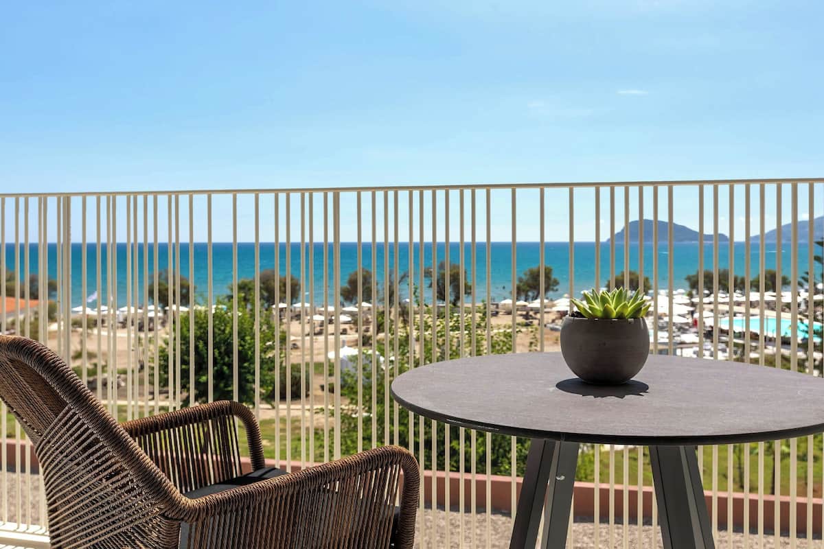A balcony with a wicker chair and a small table with a potted plant, overlooking a beach and resort area.