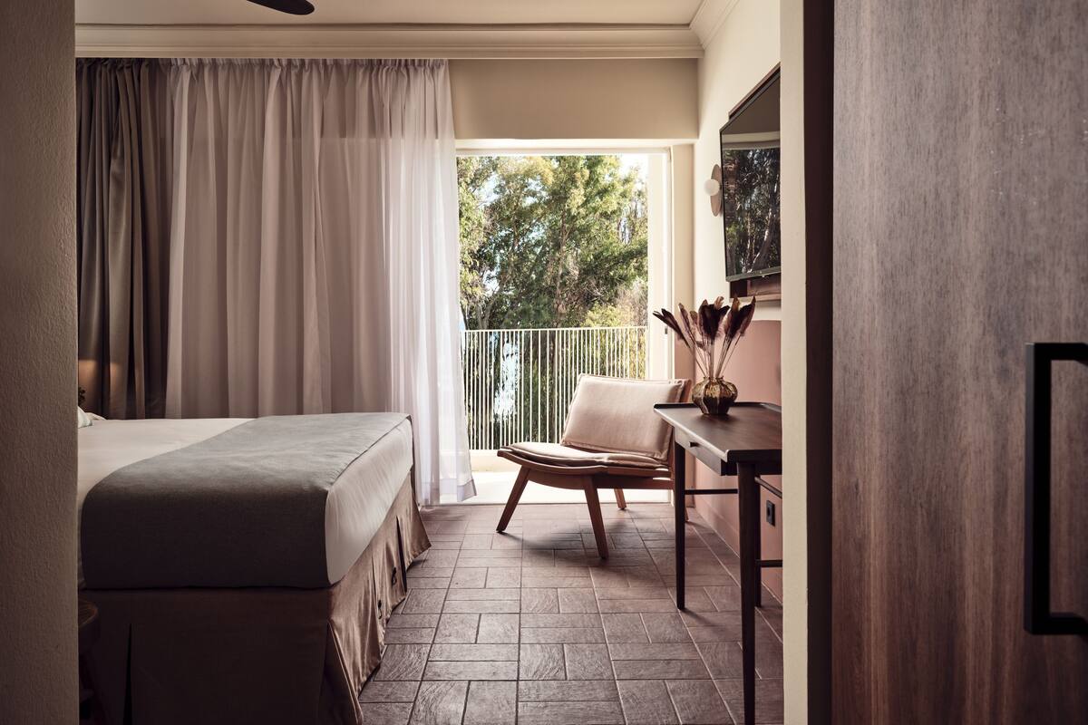 A bedroom with a bed, a chair, a table, and a balcony with a view of trees.