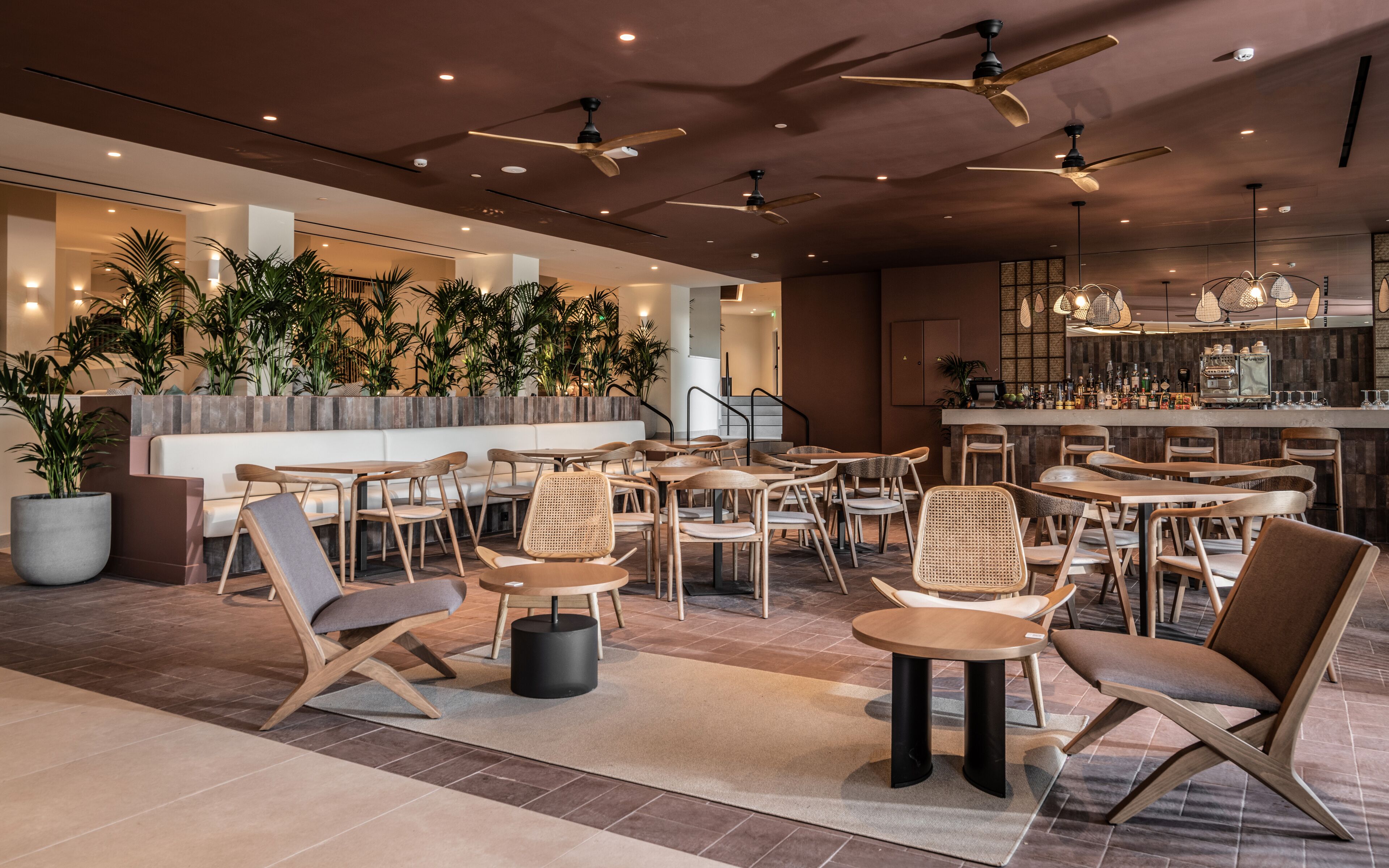 Un espacio interior moderno con varias mesas y sillas, ventiladores de techo y una zona de bar con mostrador y diversas botellas.