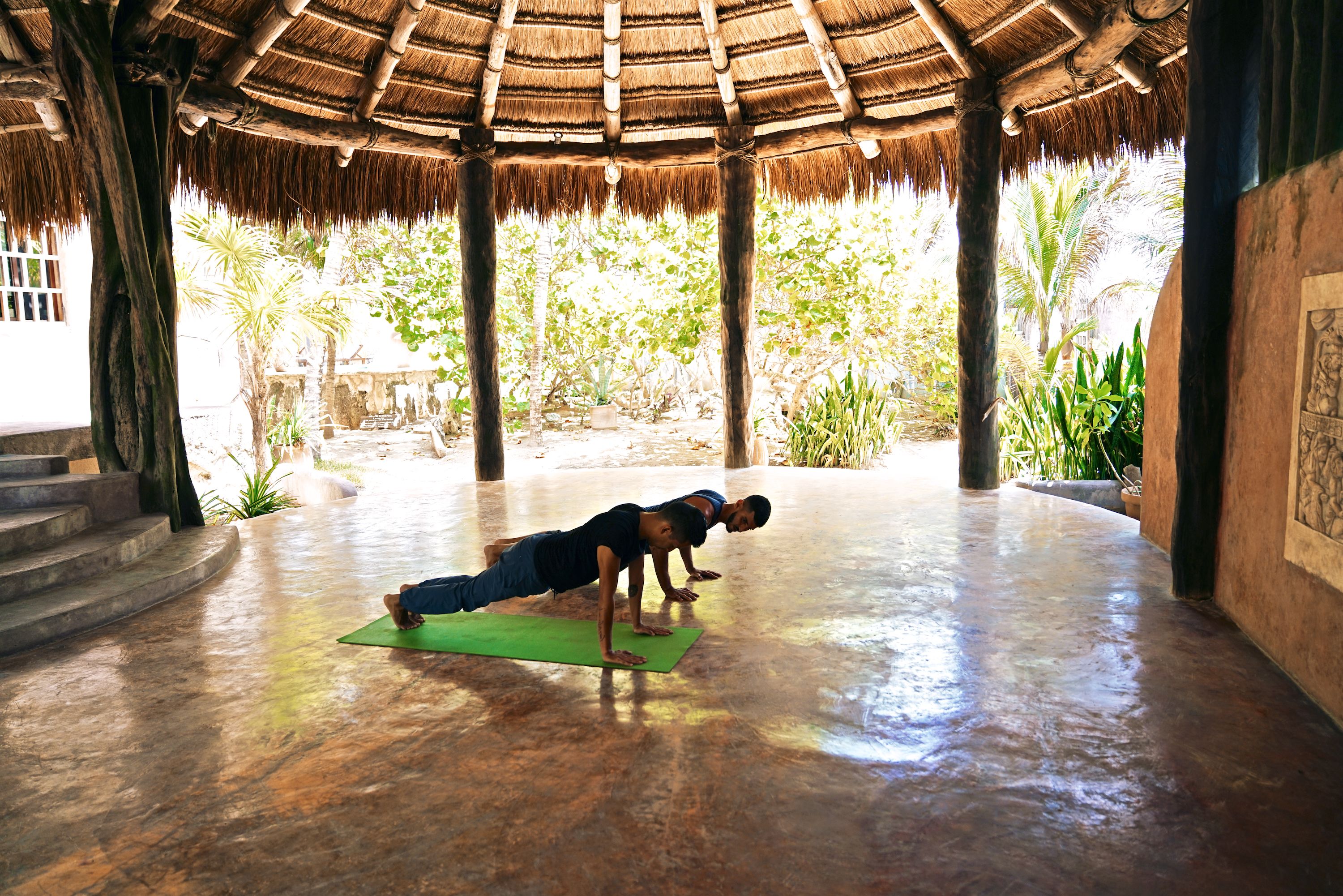 A person performing a plank exercise on a green mat in a spacious room with a thatched roof and a view of lush greenery outside.