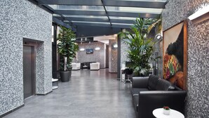 A modern lobby with a glass ceiling, tiled walls, a black leather sofa, a small round table, and potted plants. - Hotel Galileo Prague (Prague)