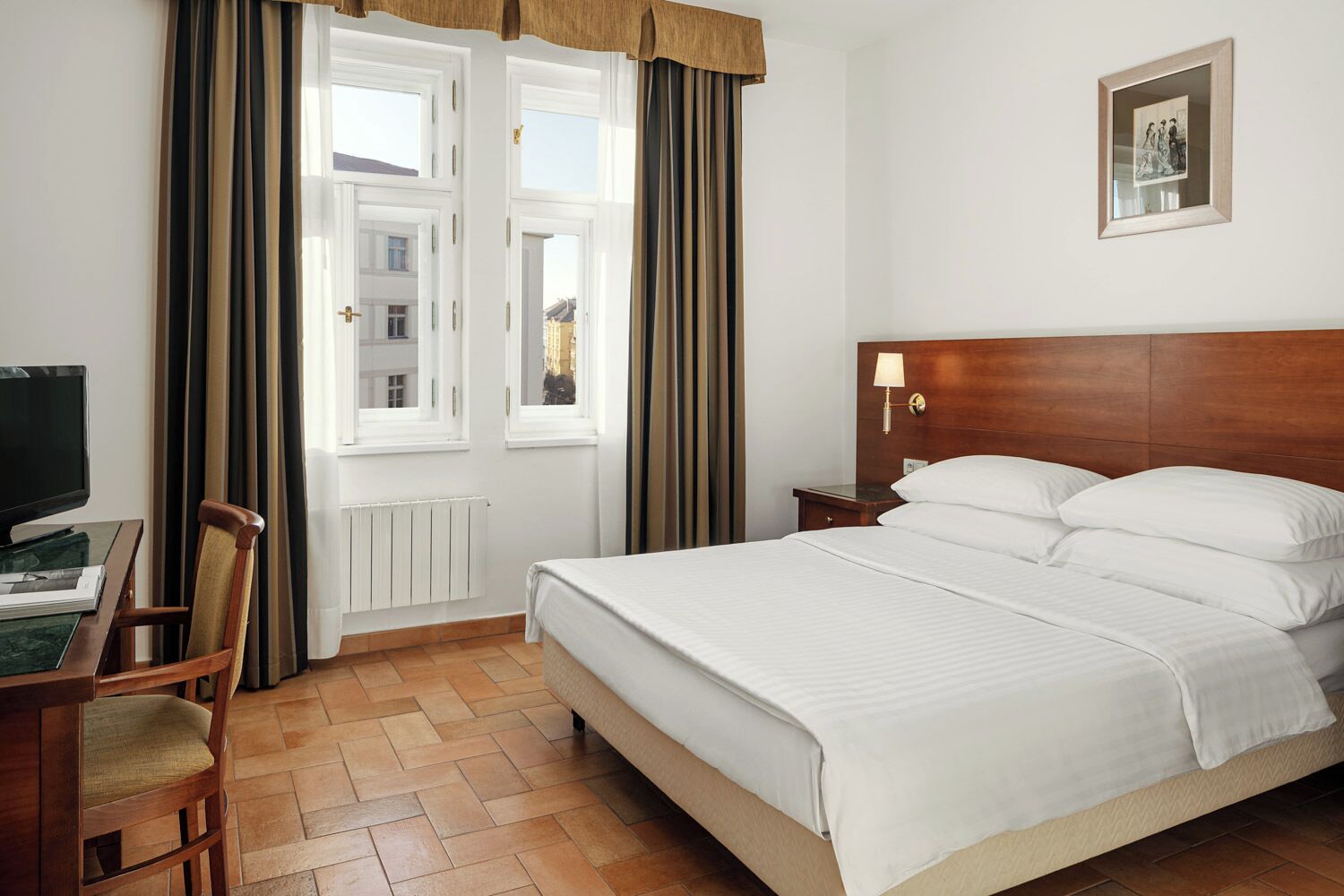 A hotel room with a bed, a desk with a computer, a chair, a nightstand, and a wall-mounted lamp.