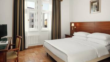 A hotel room with a bed, a desk with a computer, a chair, a nightstand, and a wall-mounted lamp.