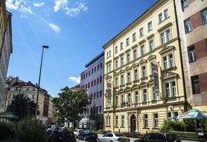 Un edificio storico dall'architettura classica, una strada con auto parcheggiate e un cielo azzurro limpido.