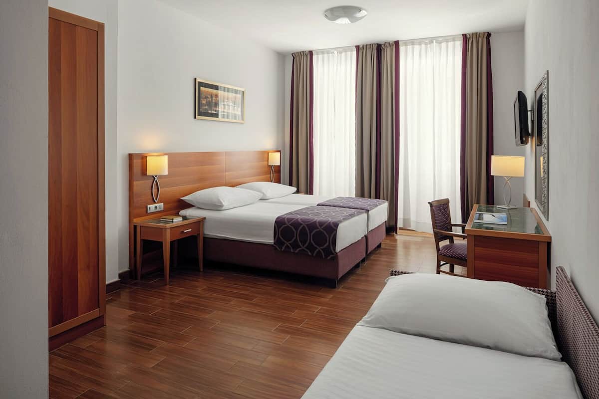A hotel room with a wooden floor, a bed with white linens, a desk with a lamp, and a chair.