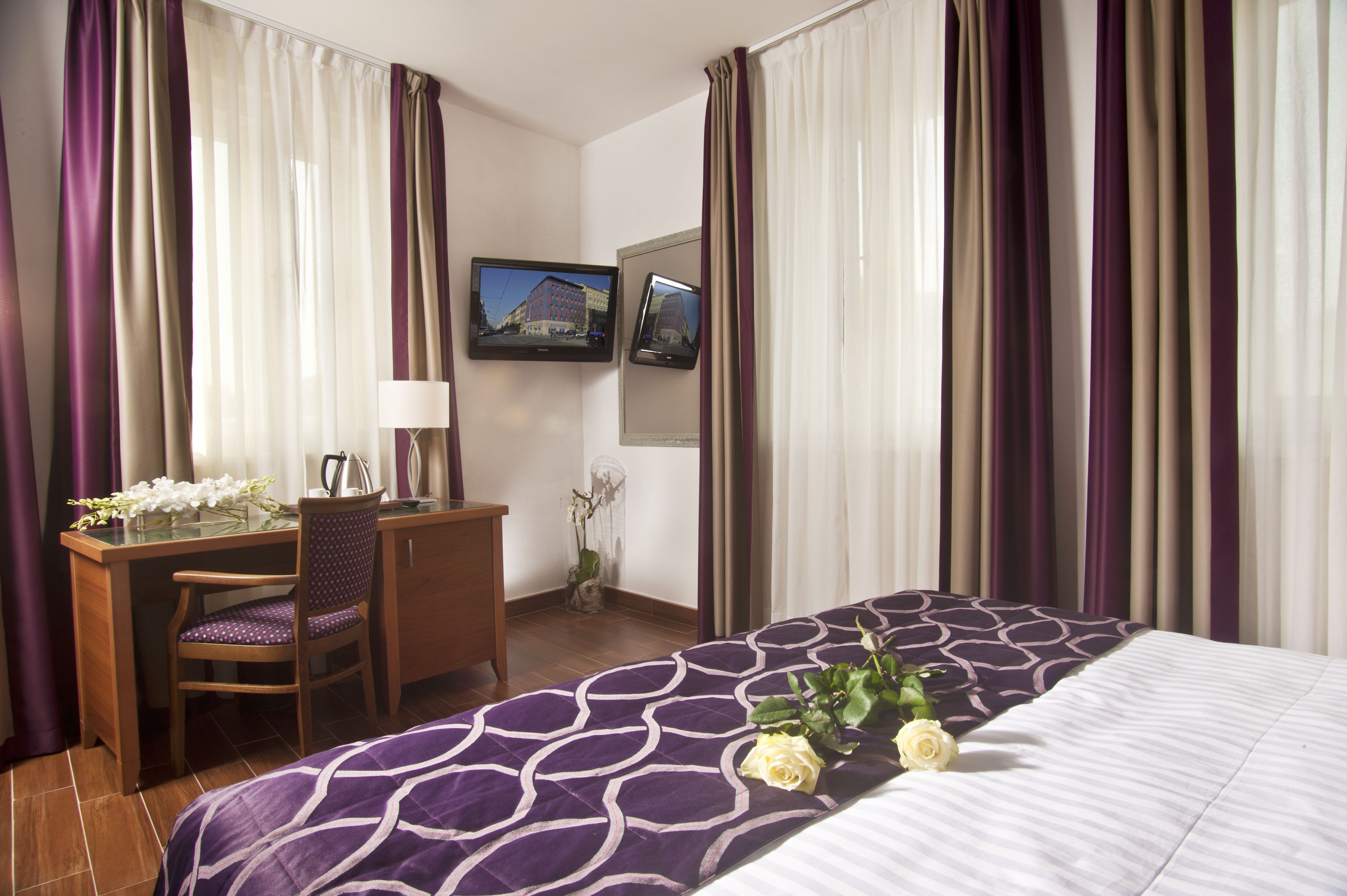 A hotel room with a bed, a desk with a chair, a television, and a vase with flowers.