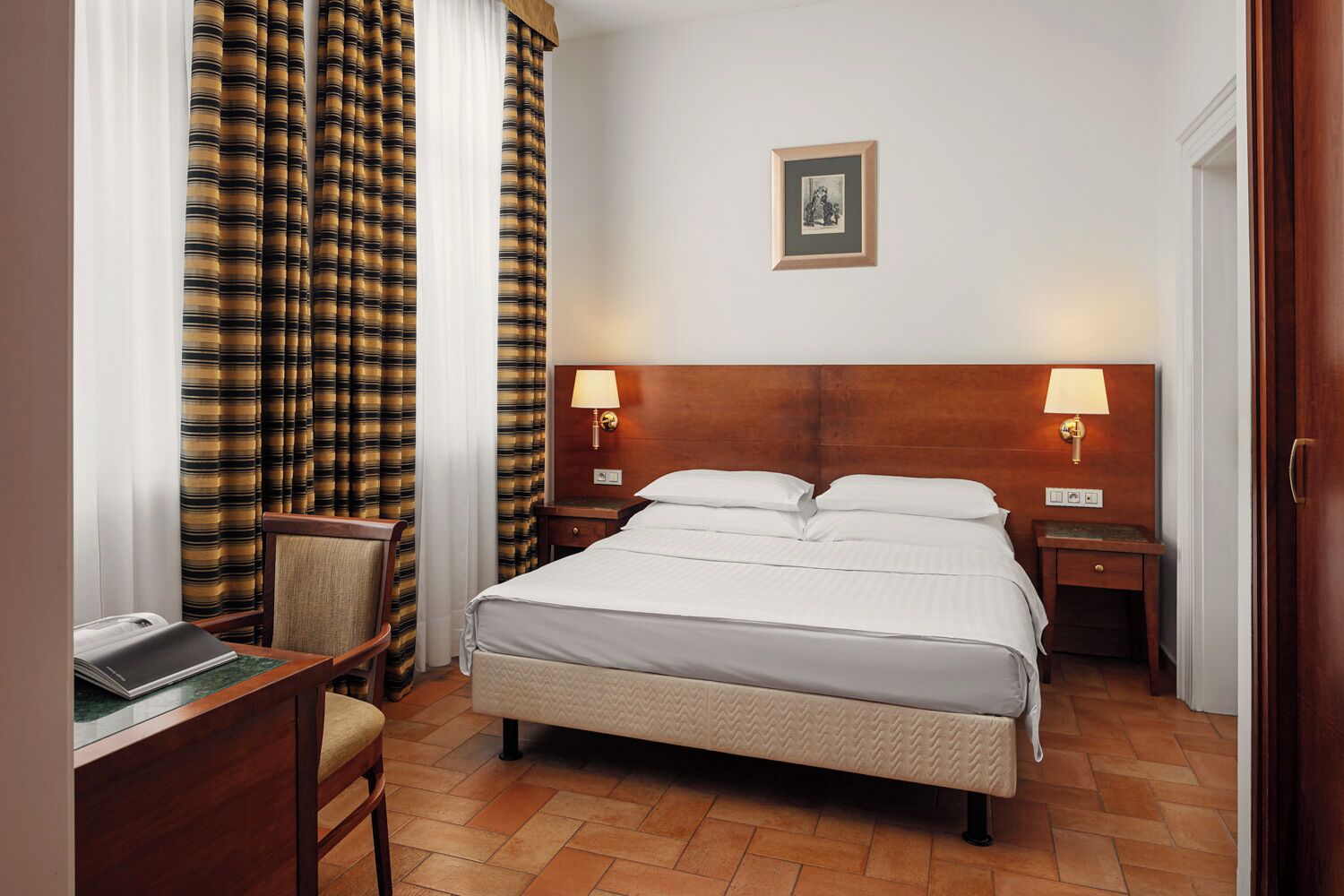Una habitación de hotel con una cama, dos mesitas de noche con lámparas, una silla, un escritorio con una computadora portátil y un cuadro en la pared.