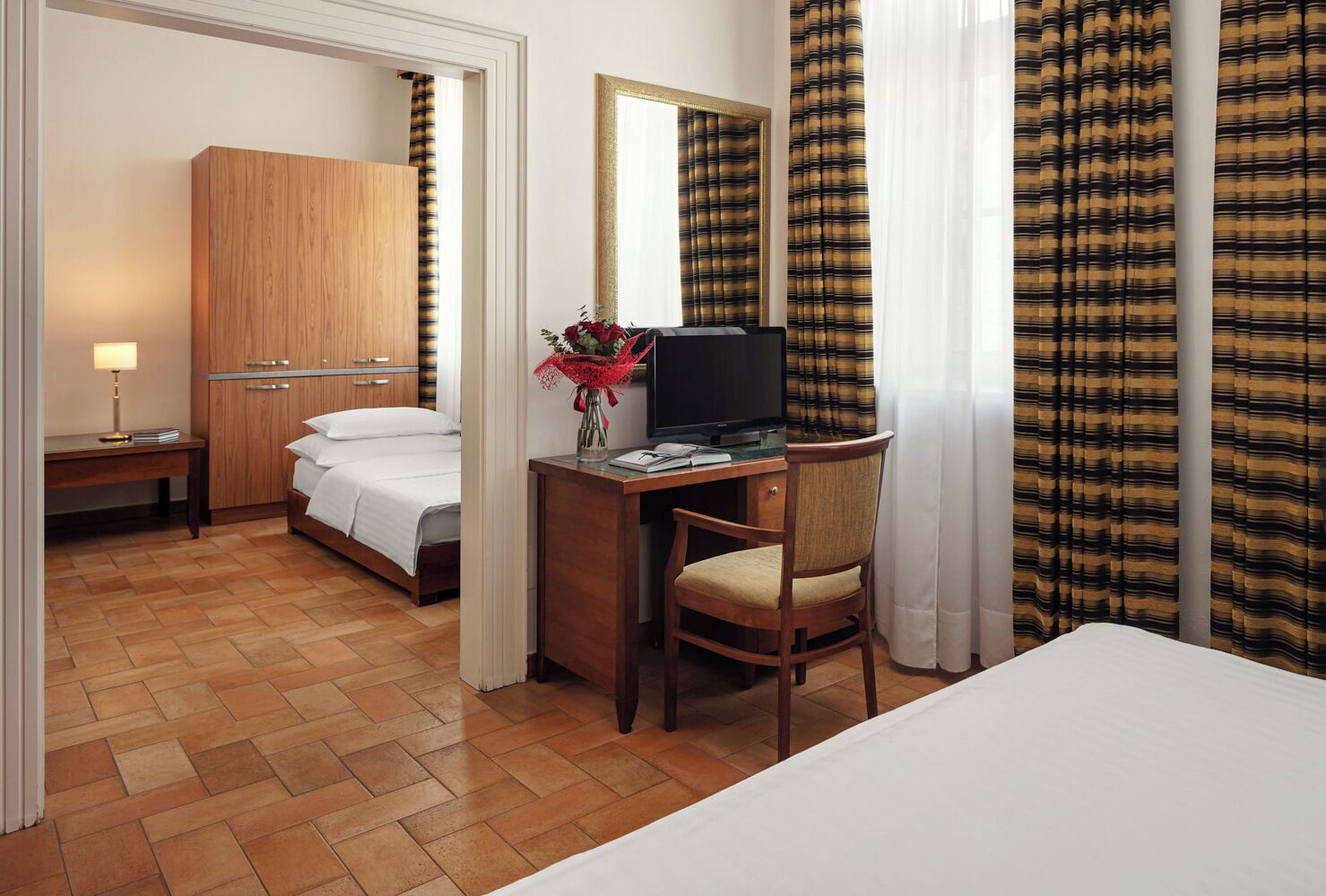 A hotel room with a bed, a desk with a computer, a chair, a TV, a bedside table with a lamp, and a wardrobe.