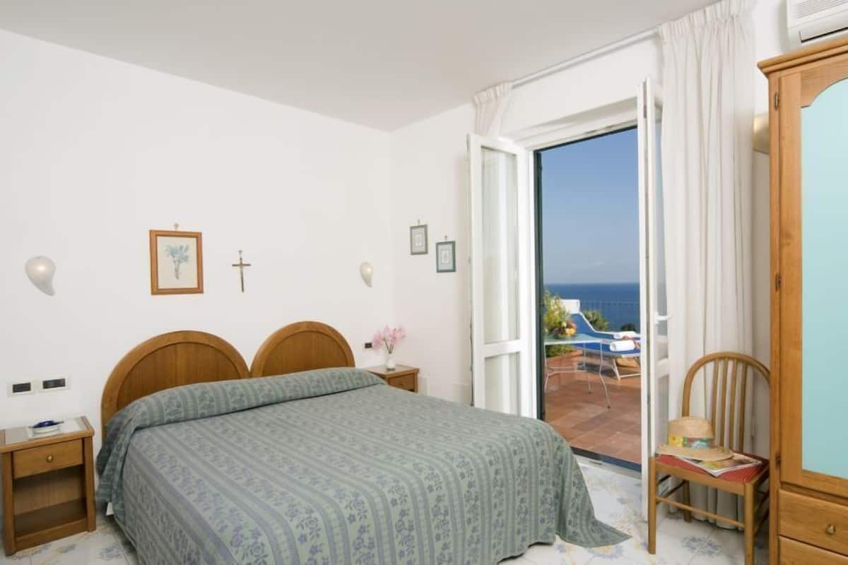 Un dormitorio con cama, una mesita de noche de madera, una silla, un armario y vista al mar a través de una puerta abierta.
