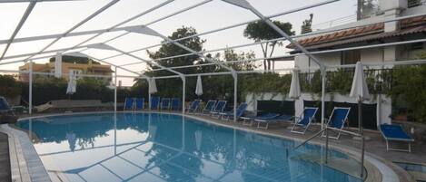 Una piscina con agua azul, rodeada de sillas de descanso y sombrillas, bajo una estructura cubierta blanca.