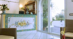 A reception area with a tiled floor, a decorative floral wall, a reception desk, wicker furniture, and a large glass door leading outside. - Hotel Gemma (Casamicciola Terme)