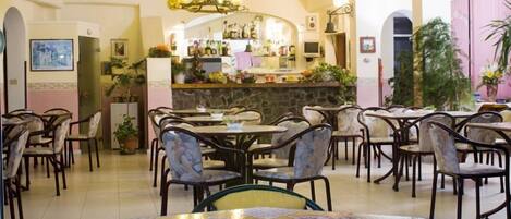 A restaurant with multiple tables and chairs, a bar area with various bottles, and potted plants.