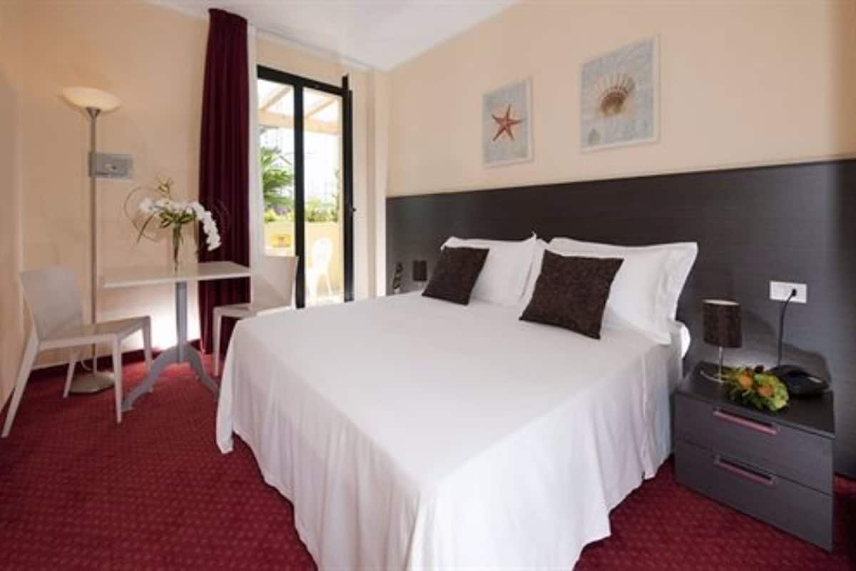 A hotel room with a large bed, a desk, and a chair. There is a window with a view of greenery outside.