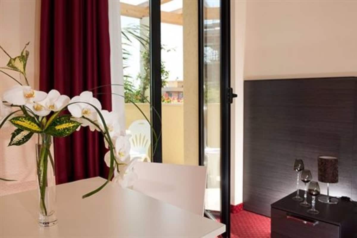 A hotel room with a dining table, a vase with white flowers, a chair, a small table with wine glasses, and a view of the outdoors.