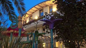 Ein Spielplatz mit Rutsche und Klettergerüsten, umgeben von Palmen und einem Gebäude mit gewölbtem Dach.