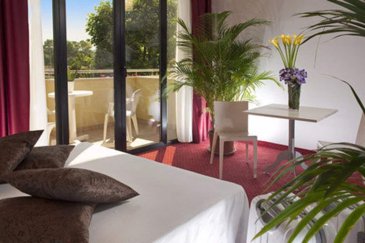 A hotel room with a bed, a small table with a vase of flowers, a chair, and a potted plant.