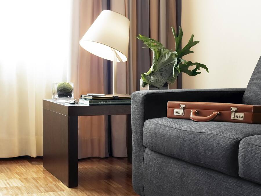 A modern hotel room with a grey sofa, a small table with a lamp, a plant, and a briefcase.