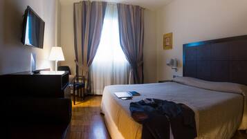 A hotel room with a bed, a desk with a lamp, a chair, a TV, and a window with curtains.
