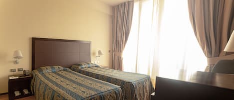 A hotel room with two beds, a desk, a chair, a lamp, and a window with curtains.