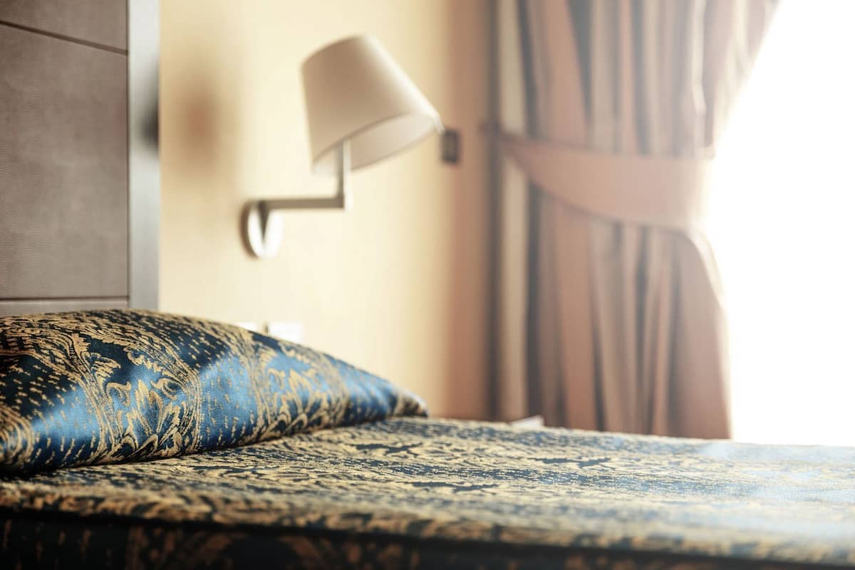 A bed with a patterned cover, a wall-mounted lamp, and a curtain.