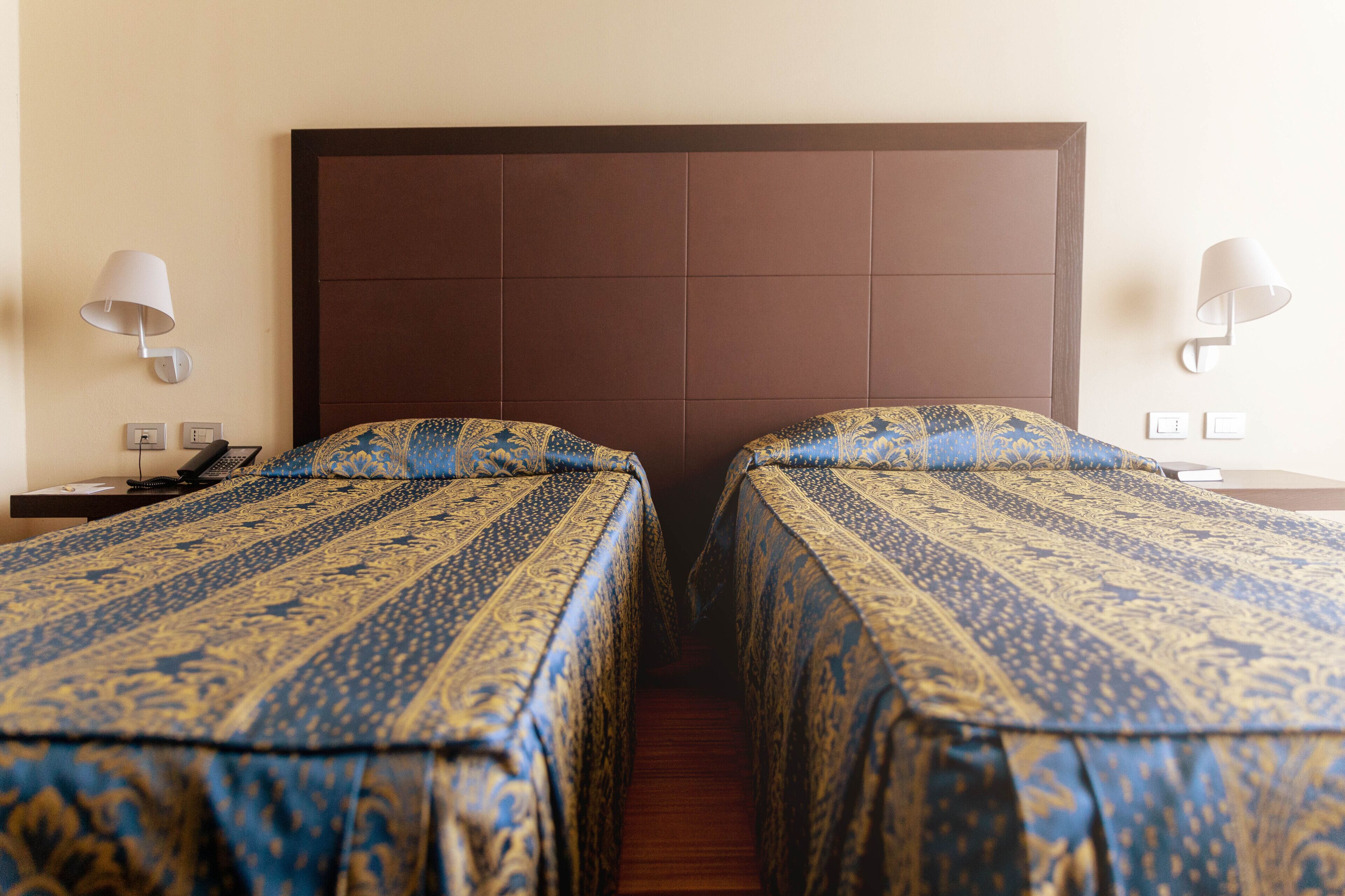 Camera d'albergo con due letti, ciascuno con una coperta a motivi decorativi, una testiera in legno, lampade da comodino e un comodino con un telefono.