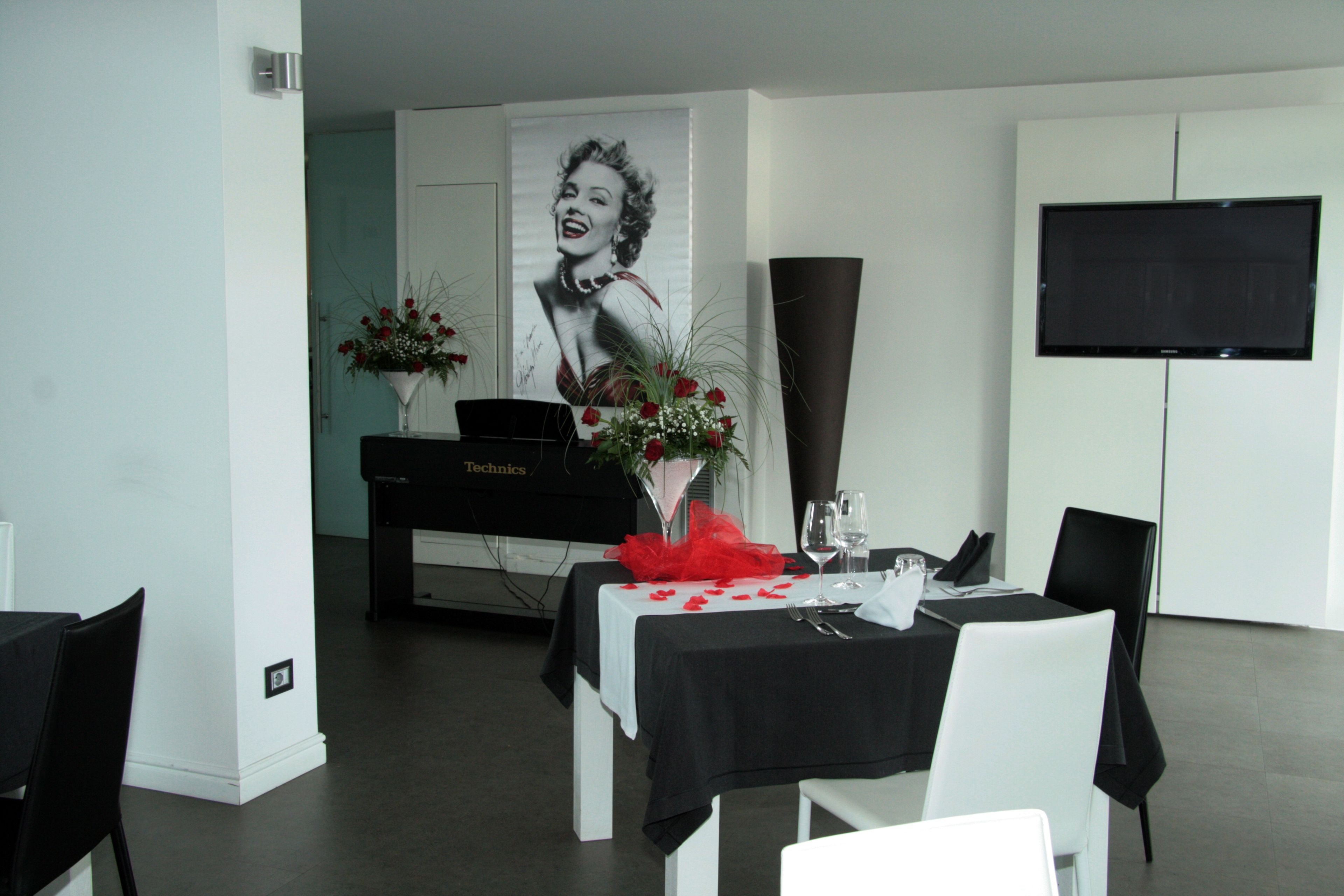 Un comedor con mantel negro, sillas blancas, un piano y un televisor grande en la pared.