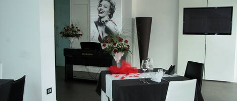 Un comedor con mantel negro, sillas blancas, un piano y un televisor grande en la pared.