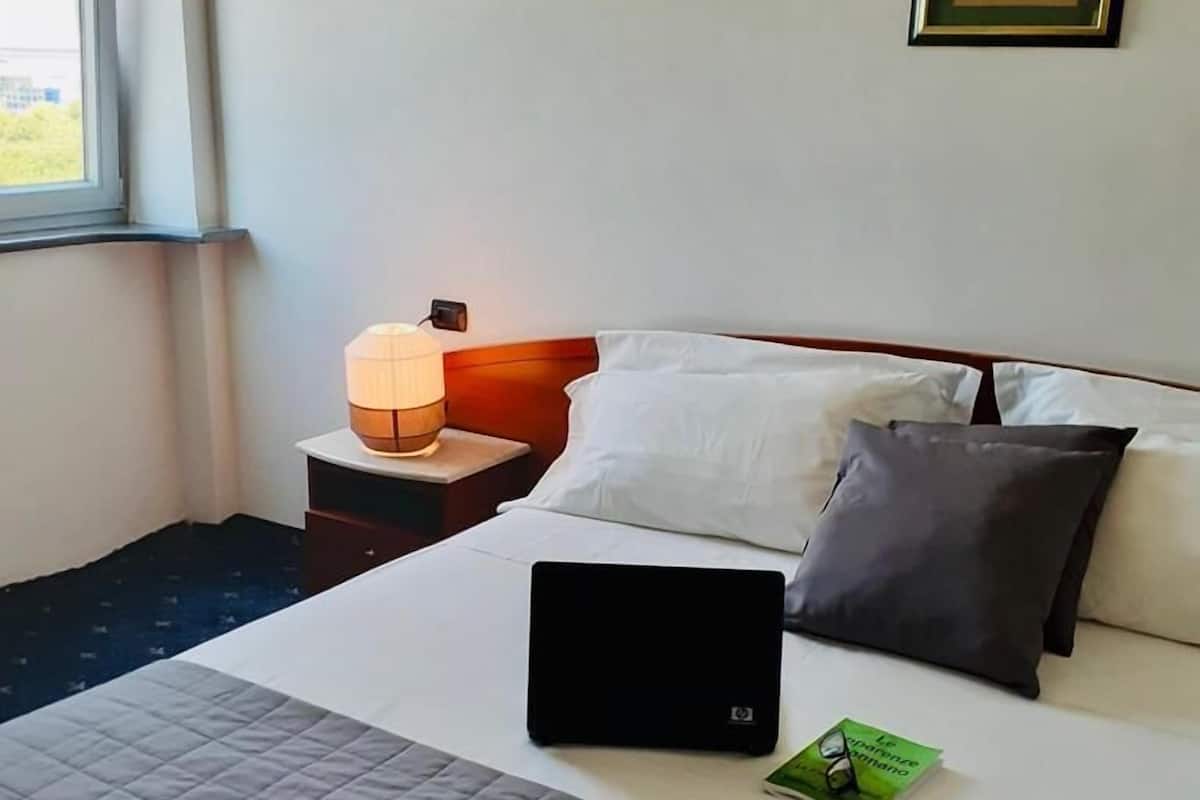 A hotel room with a bed, a bedside table with a lamp, a laptop, and a framed picture on the wall.