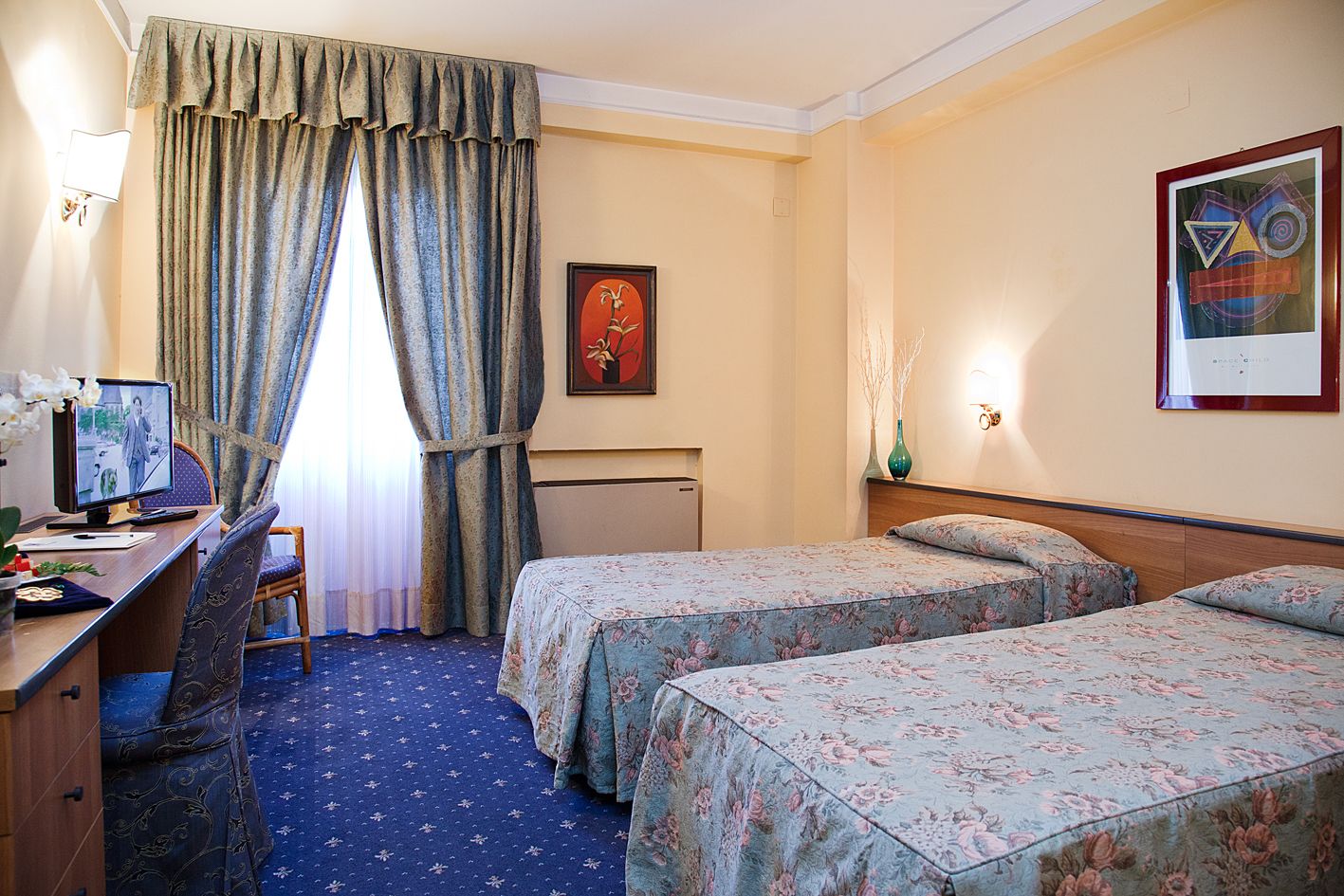 A hotel room with two beds, a desk with a computer, a chair, a desk lamp, and a painting on the wall.