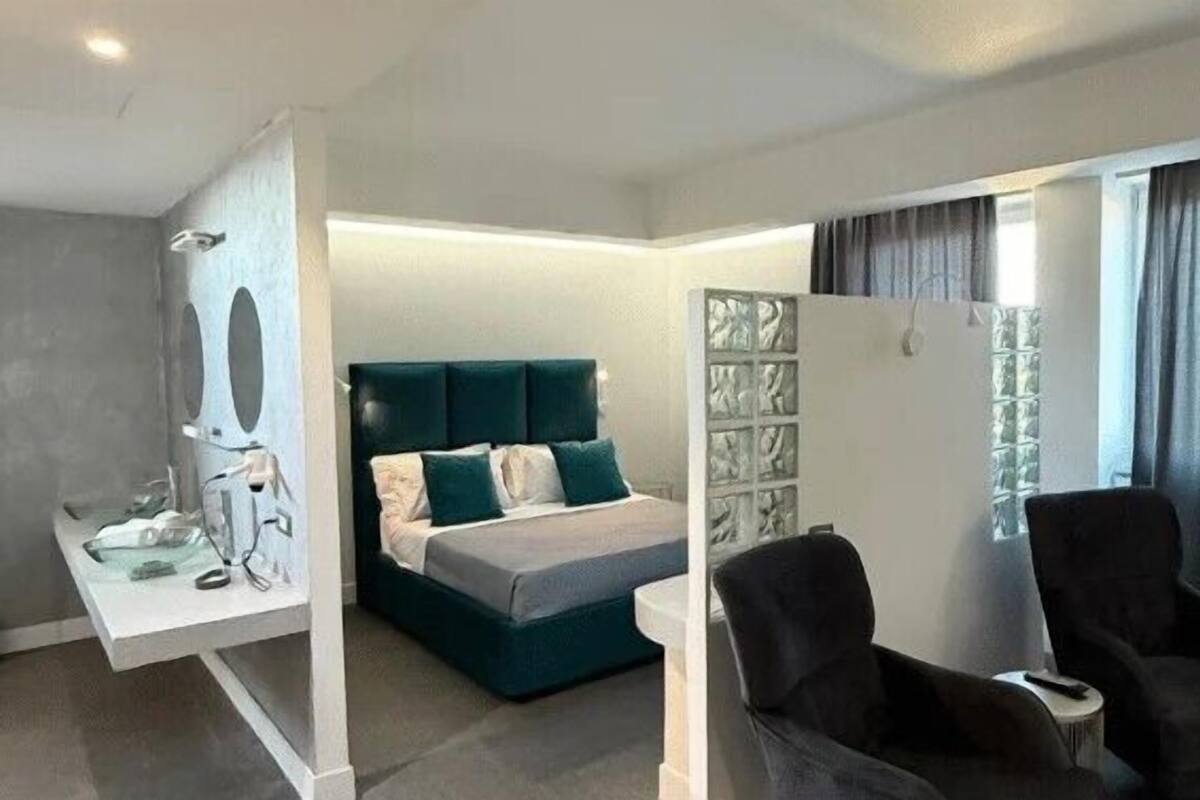 A modern hotel room with a bed, two armchairs, a bedside table, and a large window with curtains.