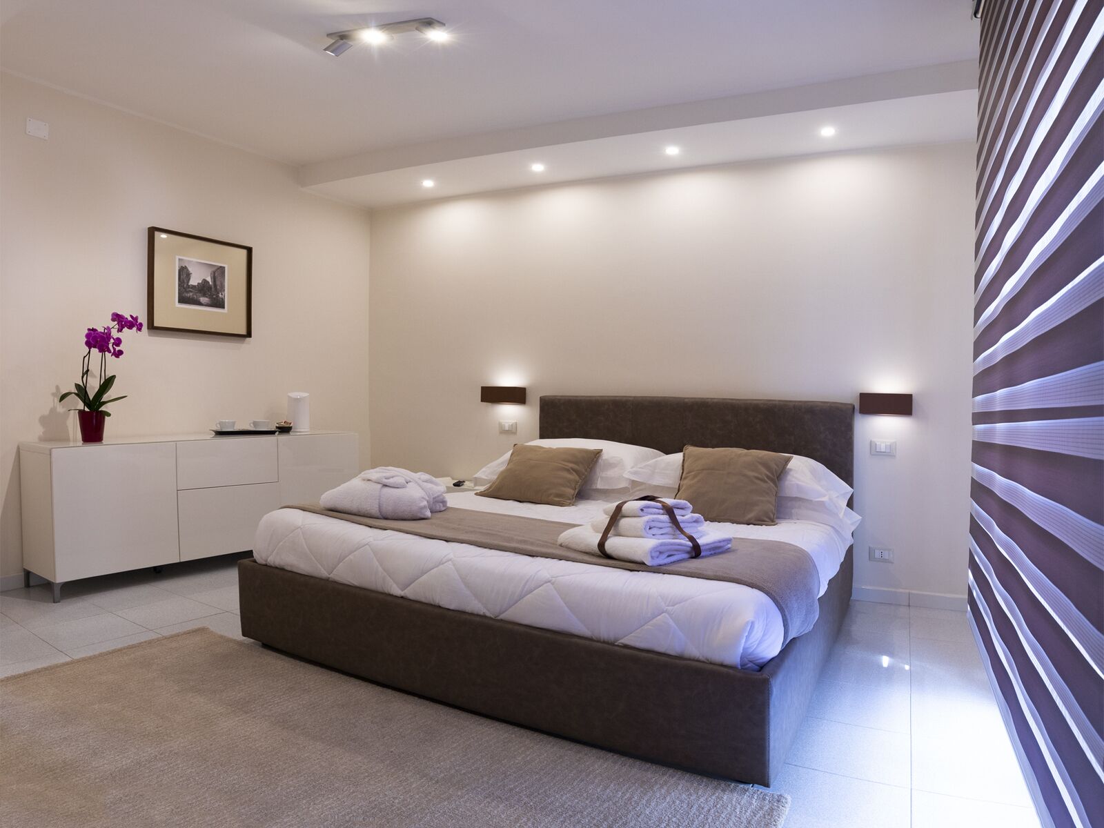 A modern bedroom with a large bed, bedside tables, a nightstand, a framed picture, and a potted plant.