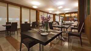 Un comedor de hotel con varias mesas y sillas, un jarrón con flores, y vista a otro espacio para comer.