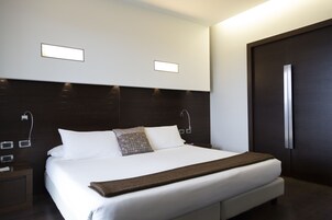 Quarto de hotel moderno com uma cama grande, cabeceira em madeira escura, mesa de cabeceira com abajur e porta com puxador.