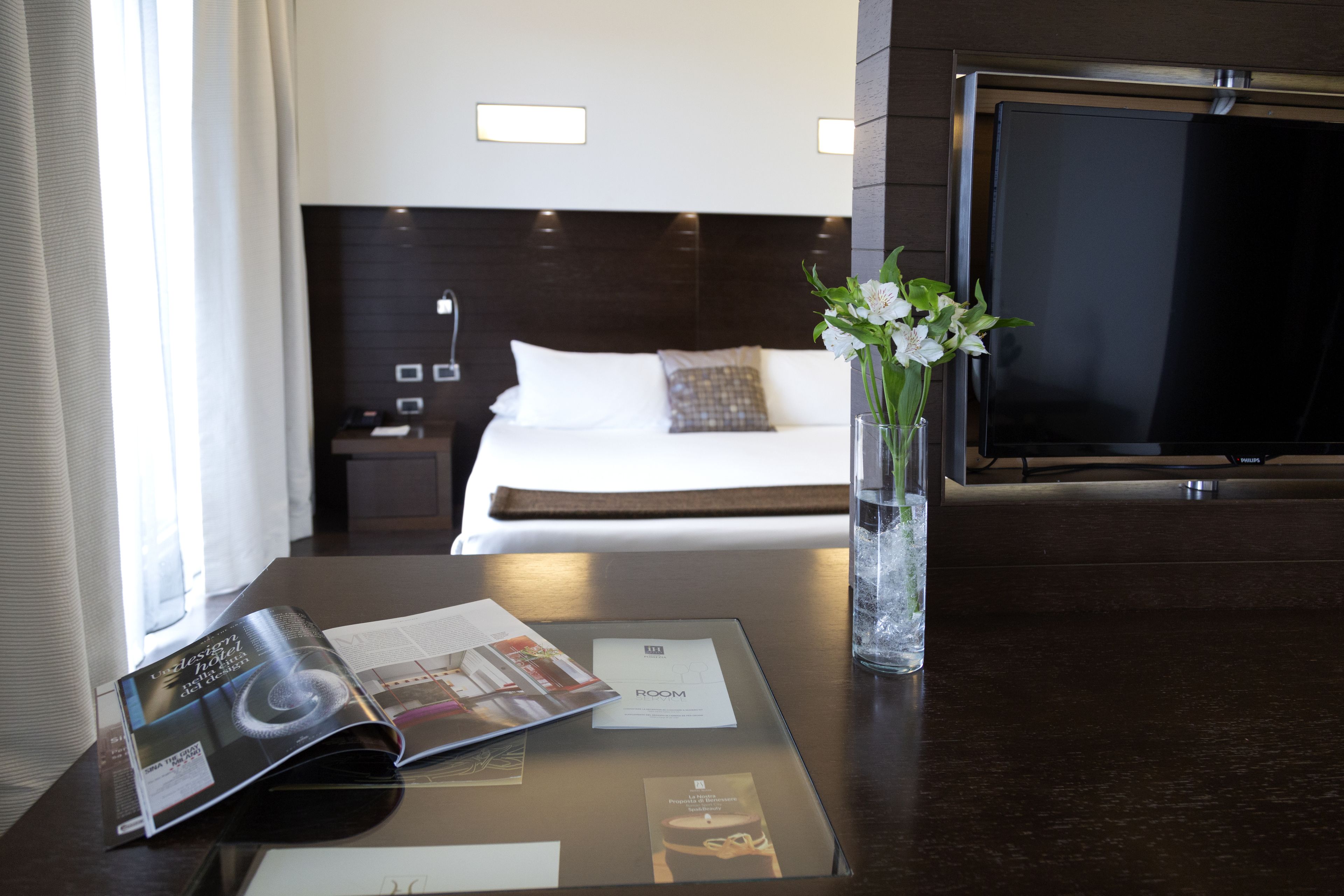 Une chambre d’hôtel avec un lit, une télévision, une table en verre avec un magazine, un vase avec des fleurs et une fenêtre avec des rideaux.