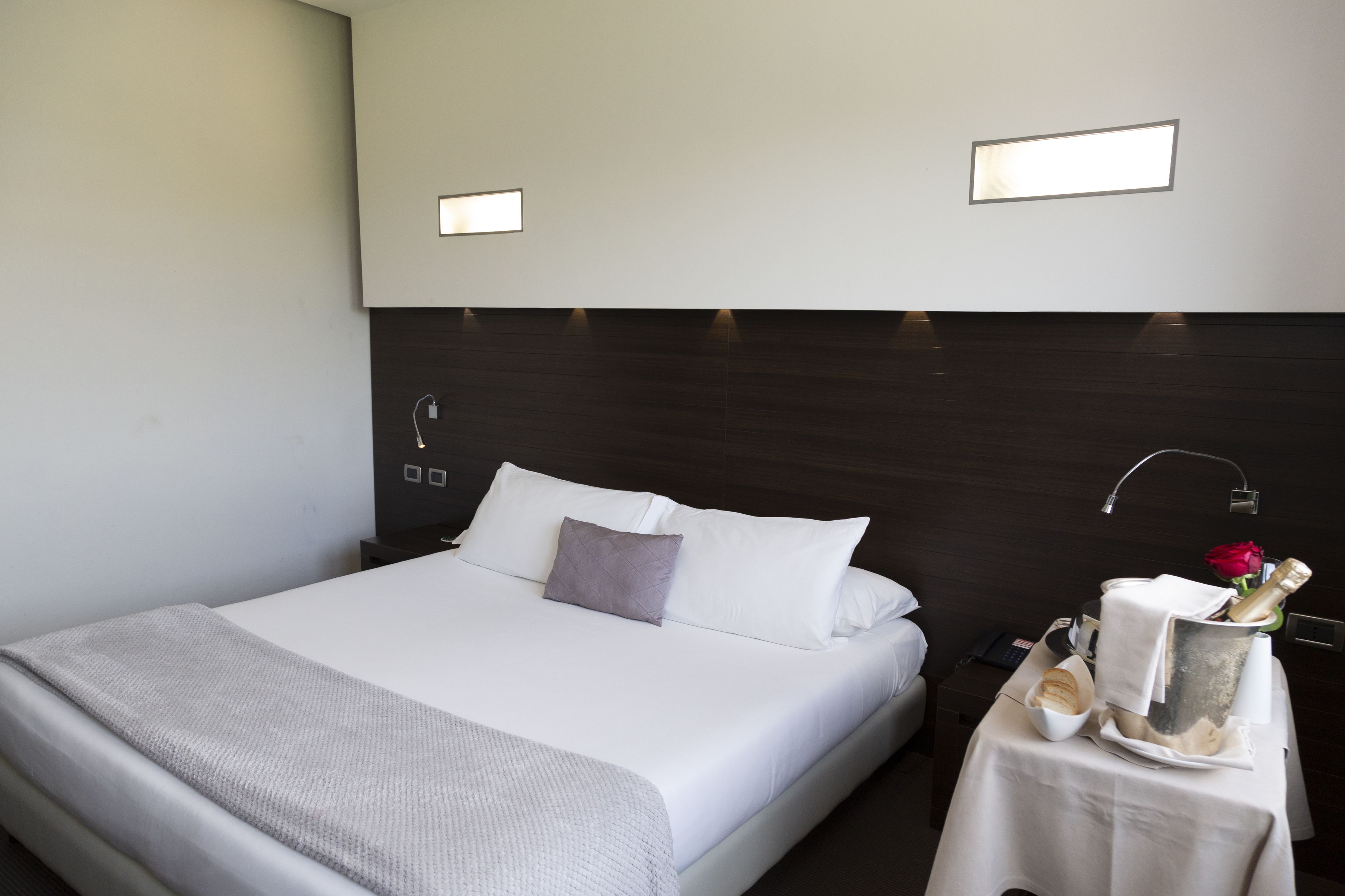 Una cama bien hecha con sábanas blancas, una almohada gris y una mesita de noche con una botella, vasos y una rosa roja.