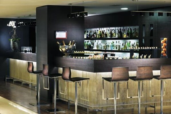 A modern bar area with a dark wood counter, multiple bar stools, and shelves stocked with bottles. - HOTEL SELENE POMEZIA (Pomezia)