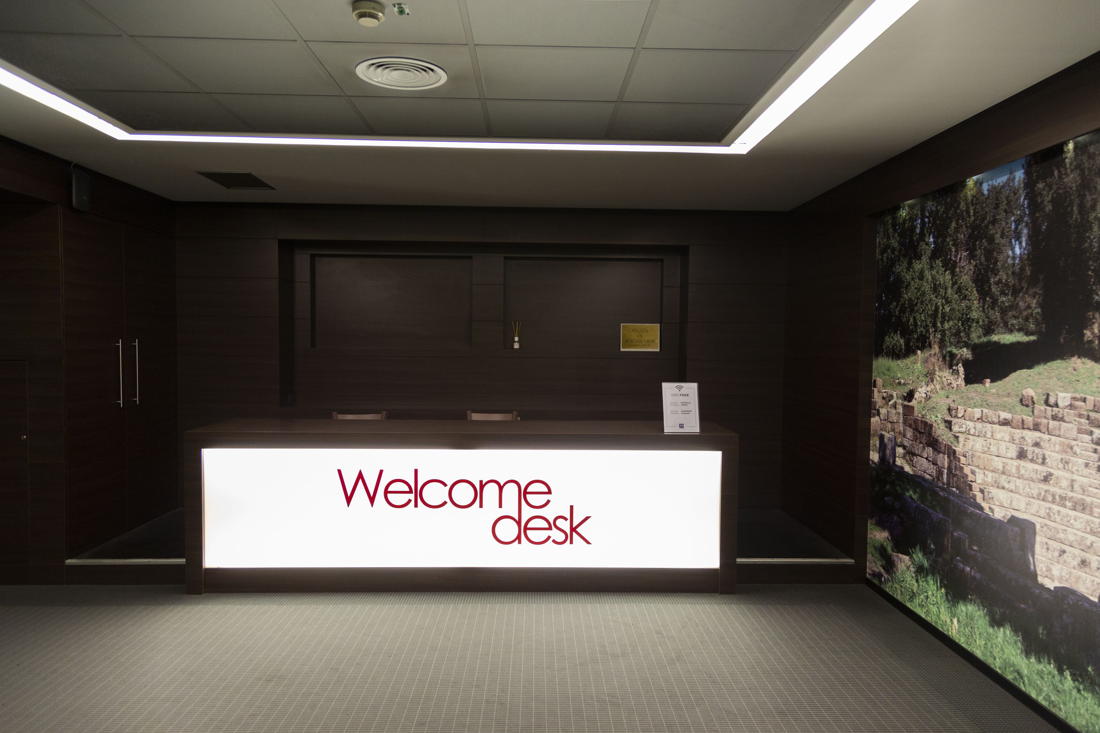 Un mostrador de recepción con un cartel que dice 'Welcome desk', una pantalla digital grande mostrando un paisaje y un piso de baldosas.
