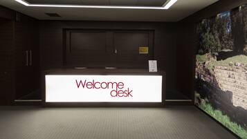 Un mostrador de recepción con un cartel que dice 'Welcome desk', una pantalla digital grande mostrando un paisaje y un piso de baldosas.