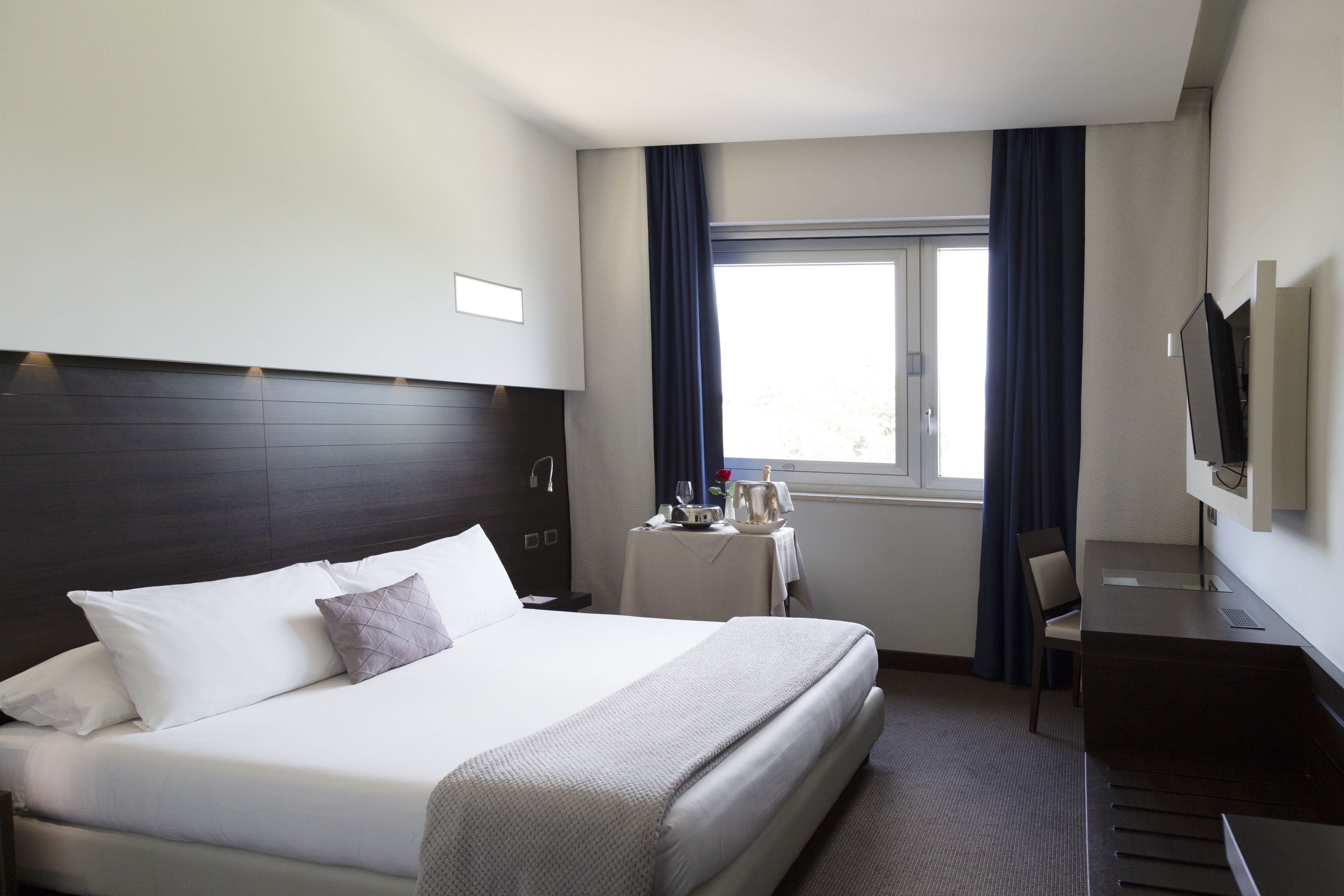 Una camera d'albergo con un letto, una scrivania, una sedia, una televisione e una finestra con tende.
