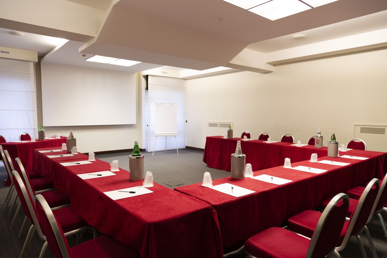 Un salón de conferencias con mesas y sillas rojas, una pizarra y una pantalla de proyector.